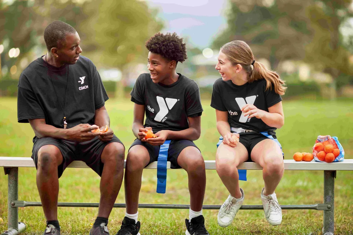 A Y coach sits with two soccer players on a bench as they snack on mandarins.